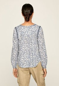 Blusa floreale bianca e blu, con maniche lunghe a sbuffo, scollo rotondo e una vestibilità morbida. Presenta una stampa floreale e una parte posteriore leggermente arricciata.