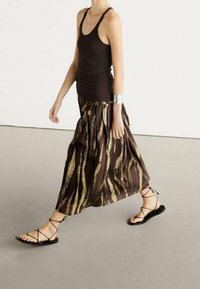 Een bruin en beige gemarmerde maxi-rok hangt van de taille; gecombineerd met een aansluitend zwart tanktop en platte zwarte sandalen. Zilveren armband om de pols.