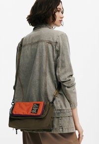 Brun och orange tvärs över kroppen väska med en dragkedjeficka, med en lila accent och "Desigual" logotyp; tillverkad av slitstarkt tyg med en slät textur.