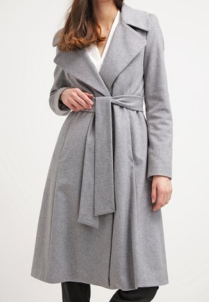Femme portant un long manteau en laine gris ceinturé avec de larges revers, par-dessus un haut blanc et un pantalon noir, sur un fond neutre.