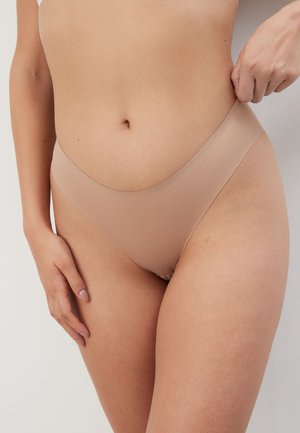 Femeie care ajustează lenjeria bej fără cusături, arătând zona mediană și partea superioară a coapselor pe un fundal simplu.