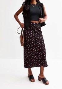 Kvinde iført sort ærmeløs vest, sort crop top, sort blomster midi-nederdel, brun skuldertaske og sorte hælede sandaler, som står foran en hvid baggrund.