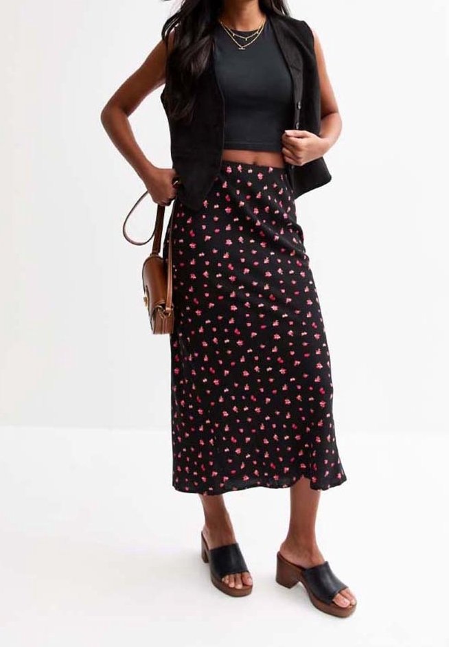 Kvinde iført sort ærmeløs vest, sort crop top, sort blomster midi-nederdel, brun skuldertaske og sorte hælede sandaler, som står foran en hvid baggrund.