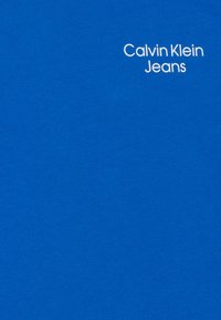 Camiseta de algodón azul brillante con el texto del logo "Calvin Klein Jeans" en blanco en una tipografía sencilla, textura suave y mangas cortas.