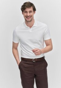 Vit kortärmad polo med krage, i kombination med en mörkbrun flätad bälte och byxor. Slät tyg med en figursydd design.