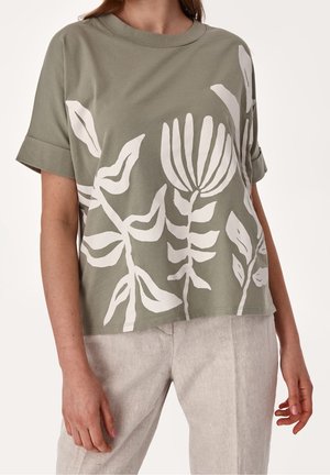 Mujer vistiendo una camiseta suelta de color verde oliva con estampados florales abstractos en blanco y pantalones de lino beige claro, de pie con los brazos relajados.