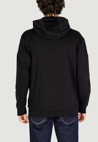 Felpa nera con cappuccio realizzata in tessuto morbido, dal taglio ampio e maniche lunghe. Cuciture visibili e una leggera trama. Indossata con jeans blu.