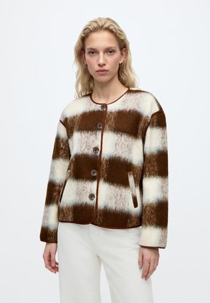 Femme aux cheveux blonds portant une veste boutonnée rayée marron et blanche et un pantalon blanc, debout devant un fond clair uni.