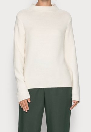 Žena nosící krémový pletený svetr s dlouhým rukávem a jemnou strukturou, doplněný tmavě zelenými volnými kalhotami, zobrazená od krku po boky.