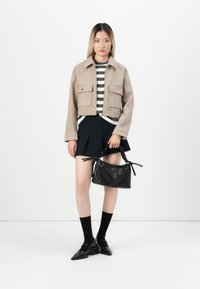 Cropped beige wool jacket with two front pockets, striped green and white shirt, black pleated skirt, black purse, and black ankle socks with shoes.