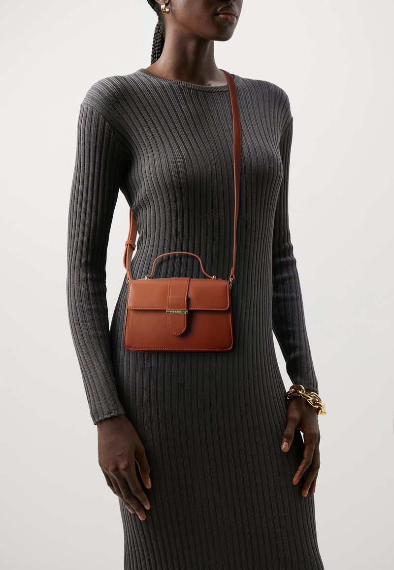 Femme portant une robe côtelée gris foncé avec un sac bandoulière en cuir marron et un bracelet chaîne en or sur un fond gris clair.