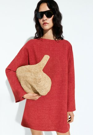 Donna con occhiali da sole neri e un vestito rosso largo, che tiene una grande borsa beige intrecciata con un manico alto.