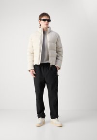 Jachetă puffer bej cu cusături verticale, hanorac gri dedesubt, pantaloni negri și adidași crem. Persoana poartă ochelari de soare negri.