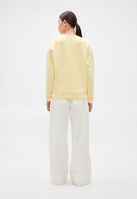 Sárga pulóver laza szabással, kerek nyakkivágással és bordázott mandzsettákkal, lazán illeszkedő fehér nadrággal párosítva. Látható minták vagy logók nélkül.