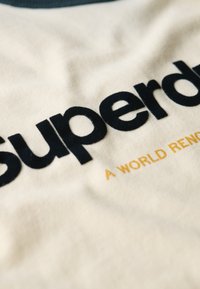 Camiseta de algodón con base crema, letras en azul oscuro "superdry" y acento dorado "UN MUNDO RENOMBRADO" en un corte relajado.