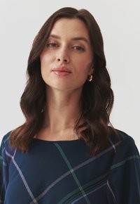 Haut bleu marine à encolure carrée, présentant un motif écossais en vert et blanc. Tissu doux avec un léger éclat ; le modèle a les cheveux ondulés.