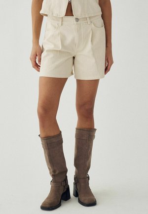 Nő bézs rövidnadrágban és hozzáillő mellényben, barna térdig érő magas sarkú csizmával, egyszerű világos háttér előtt állva.