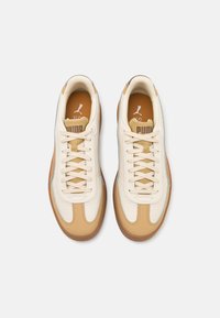 Pair of beige and white Puma sneakers with gum soles, beige laces, and branding on tongue and insole, viewed from above.