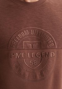 Smeđa majica s kapuljačom koja se ističe teksturiranim materijalom, rebrastim rubovima oko izreza i otiskanim grafičkim detaljima s natpisima "Freedom Unlimited" i "PME Legend".