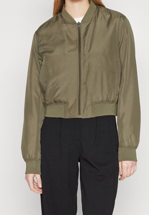 Femme portant une veste bomber vert olive avec fermeture éclair à l'avant et poignets côtelés, associée à un pantalon noir taille haute.