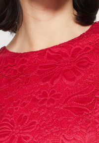 Röd spetsblus med intrikata blommönster och en slät textur, med rund halsringning och korta ärmar.