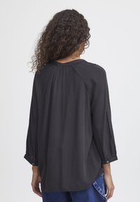 Black long-sleeve shirt with a relaxed fit, featuring subtle texture, a round neckline, and buttoned cuffs. Back pleating adds detail.