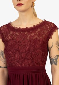 Abito borgogna con bustier in pizzo floreale con bordi scallop, una cintura larga e una gonna fluida. Il motivo del pizzo dettagliato è prominente.
