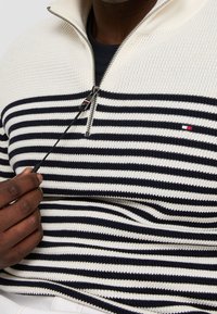 Main tenant une fermeture éclair d'un pull en tricot rayé marine et blanc avec une demi-fermeture éclair et un petit logo rectangulaire sur la poitrine.