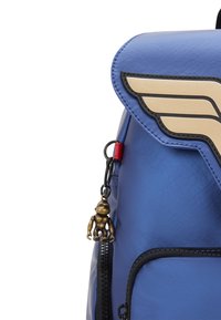 Blå syntetisk ryggsäck med en flap som har ett guldvingedesign, en dragkedjesnurra formad som en liten figur, och svarta accentstygn.