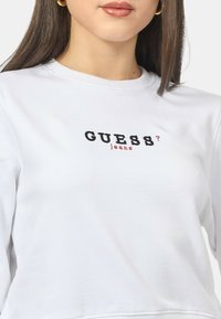 Felpa bianca con collo rotondo, caratterizzata da testo nero e rosso che dice "GUESS? jeans." Tessuto liscio e vestibilità comoda.