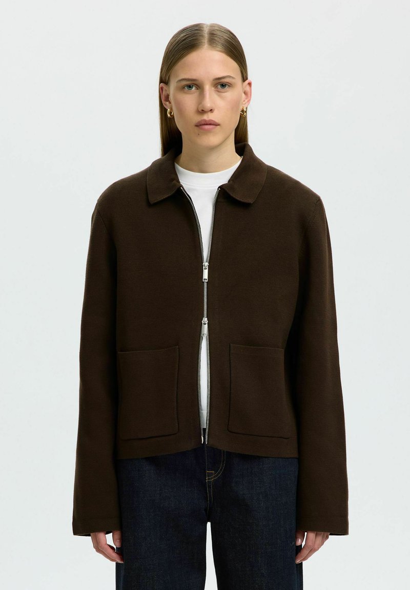 Jeune personne aux cheveux lisses porte une veste zippée marron foncé sur une chemise blanche et un jean bleu foncé, debout devant un fond uni.