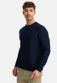 Maglione a maniche lunghe blu navy con scollatura rotonda, polsini a coste e un motivo di maglia testurizzato. Design aderente con cuciture laterali visibili.