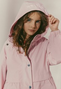 Giacca impermeabile rosa con cappuccio, dotata di bottoni a pressione e vita regolabile. Il tessuto è liscio e presenta una leggera lucentezza.