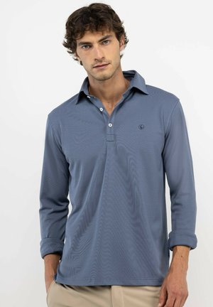 Hombre con cabello rizado castaño, que lleva una camisa polo azul de manga larga con dos botones abiertos y pantalones beige, de pie frente a un fondo blanco liso.