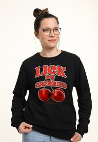 Sudadera negra con un diseño gráfico colorido que incluye el texto "LICK my CHERRIES" y dos cerezas rojas. Material de algodón, cuello redondo, mangas largas.