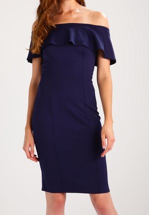 Donna che indossa un vestito aderente blu scuro, scollo a barca, lunghezza al ginocchio, con dettaglio a balze sul petto.