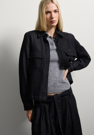 Giacca in denim nera con chiusura a bottoni e tasche, indossata sopra un maglione grigio chiaro con orlo a coste. Il modello si posa su uno sfondo neutro.