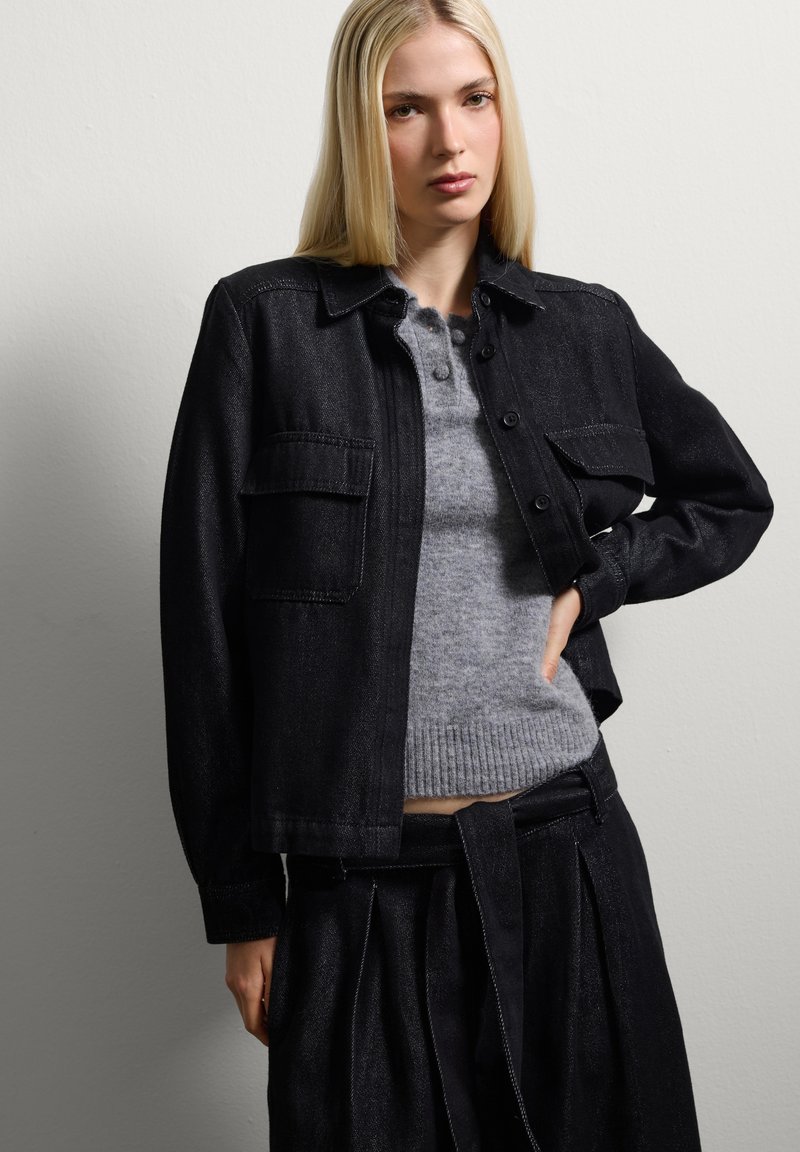 Giacca in denim nera con chiusura a bottoni e tasche, indossata sopra un maglione grigio chiaro con orlo a coste. Il modello si posa su uno sfondo neutro.