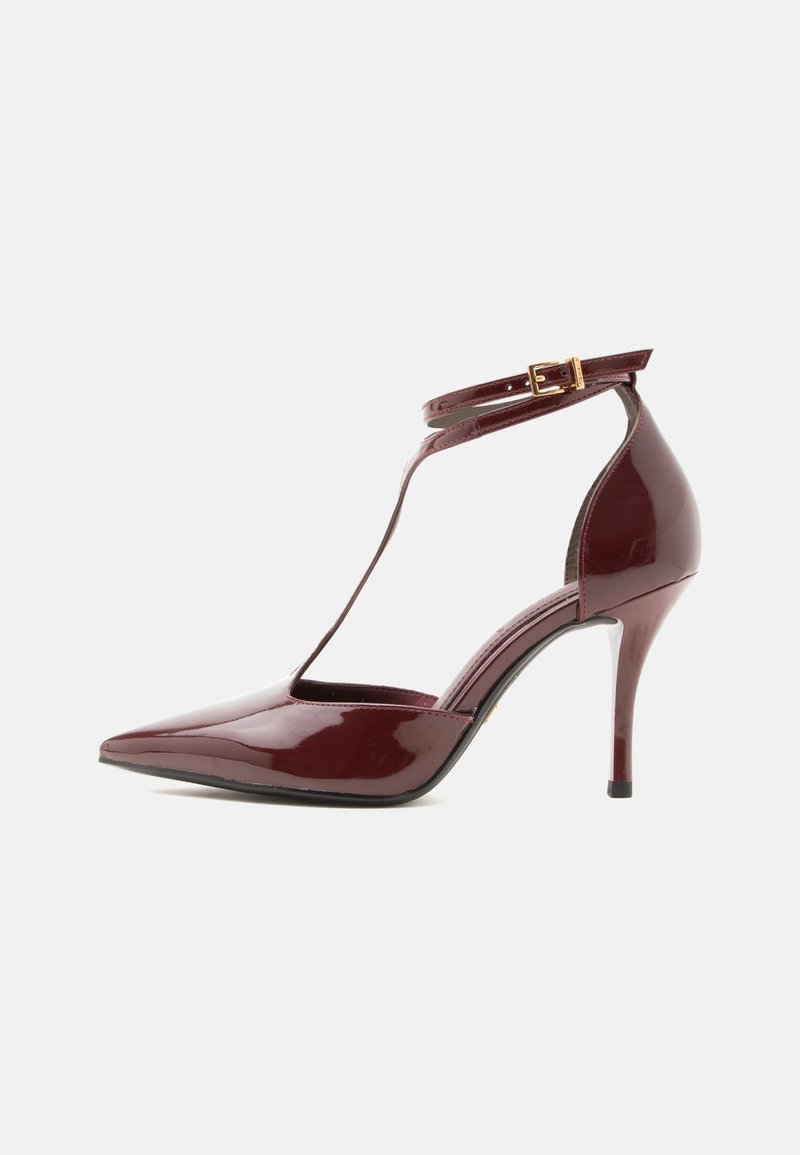Chaussure à talon haut rouge foncé brillante avec bout pointu et fine bride à la cheville fermée par une petite boucle dorée sur fond blanc.