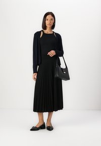 Jupe plissée noire, haut noir, cardigan bleu marine et sac à main noir. Le mannequin porte des talons noirs avec des embellissements. Textures lisses et design classique.