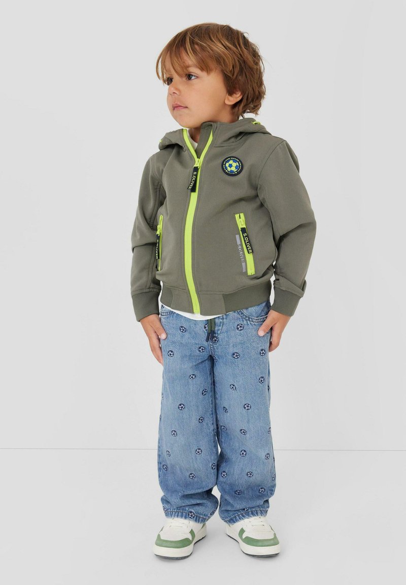 Junge steht, trägt eine grüne Kapuzenjacke mit neonfarbenen Reißverschlüssen, blaue Jeans mit kleinen Fußballmotiven und weiße Turnschuhe.