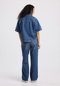 Outfit in denim con una camicia a maniche corte e pantaloni a gamba larga di un solido colore blu scuro. Il materiale presenta una texture liscia con cuciture visibili.