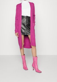 Langer rosa Cardigan, weißer Rollkragenpullover, Minirock aus schwarzem Leder und glänzende rosa Stiefeletten mit quadrater Zehenform und Reißverschlussdetails.