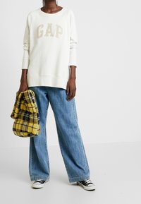 Sudadera blanca con "GAP" en letras beige, combinada con jeans de pierna ancha de color azul claro y una bolsa de cuadros amarillos. Zapatillas negras de corte bajo.
