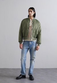 Chaqueta bomber verde oliva con cuello de canalé, camisa crema clara debajo, jeans azul claro y zapatos negros de cordones. Color sólido con detalles mínimos.