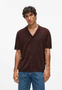 Rudas trumpas rankovėmis marškiniai su mygtukų apykakle, lengvai tekstūruotas audinys, priekiniai mygtukai ir iškaltas logotipas kairiajame krūtinės pusėje.