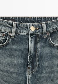 Džínové kalhoty v bledě modré barvě s mosazným knoflíkem, kontrastním prošíváním a designem s pěti kapsami. Textura je hladká s mírným opotřebením.