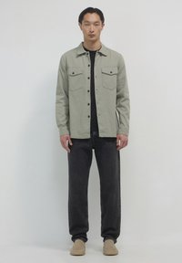 Camicia button-up in tessuto verde chiaro, con due tasche sul petto, abbinata a pantaloni in denim scuro e scarpe slip-on color cammello. Sfondo neutro.