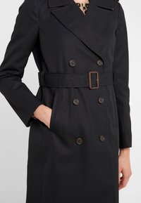 Žena oblečená v černém dvouřadém trenčkotě s páskem a rukou v kapse, na krku viditelný leopardí vzor.