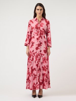 Frau trägt ein langärmeliges, rosa Maxikleid mit roten Blumenmustern, durchgeknöpft vorne, und schwarze spitze Pumps, steht vor einem einfarbigen Hintergrund.